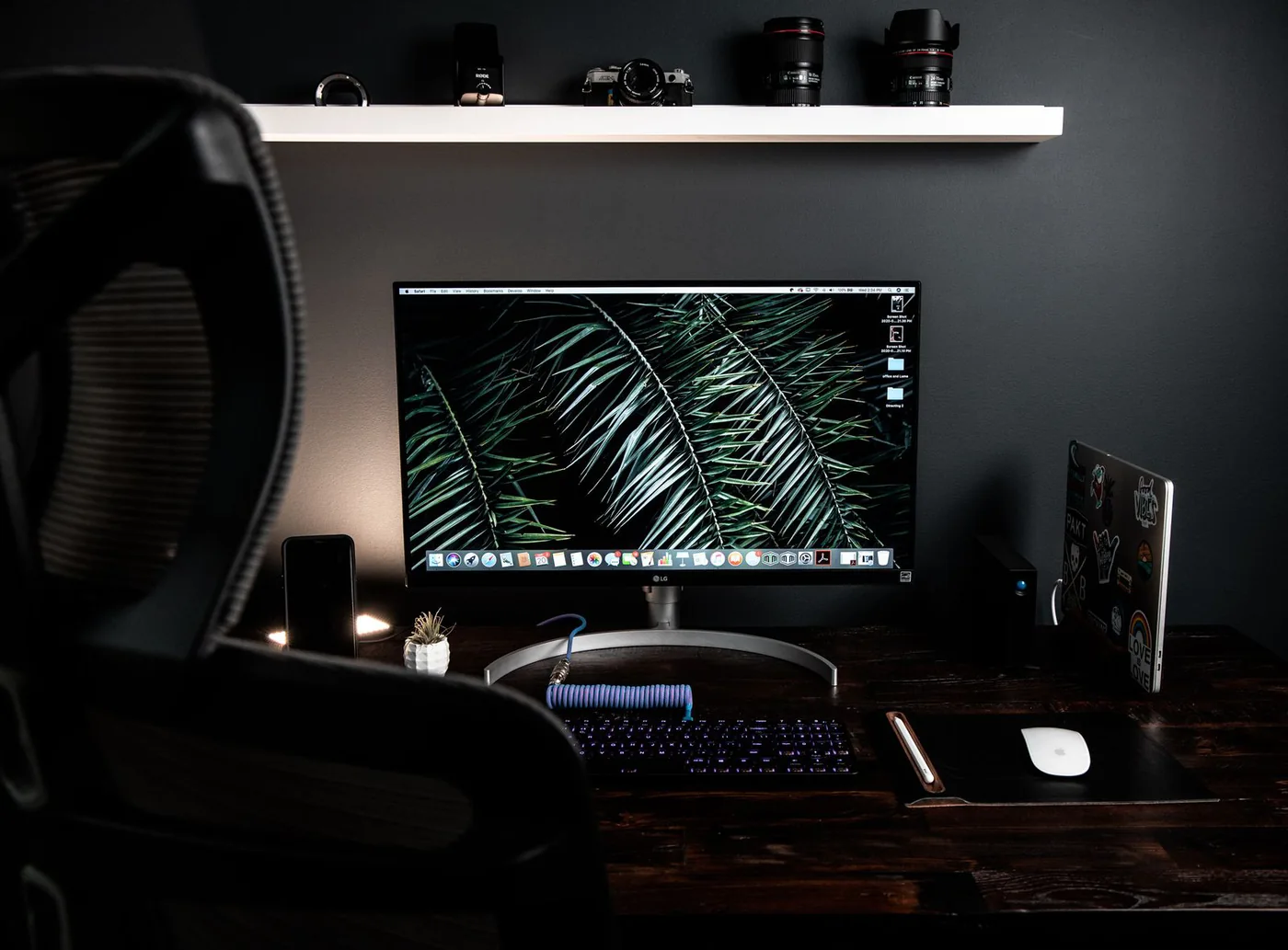 Monitor, keyboard and accessories on a dark desk below a white shelf